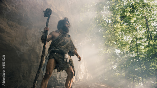 Primeval Caveman Wearing Animal Skin Holds Stone Hammer Stands Near Cave and Looks Around Prehistoric Landscape, Ready to Hunt Animal Prey. Neanderthal Going Hunting into Jungle. Low Angle Shot photo