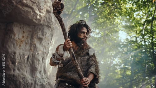 Primeval Caveman Wearing Animal Skin Holds Stone Tipped Hammer Comes out of the Cave and Looks Around Prehistoric Landscape, Ready to Hunt Animal Prey. Neanderthal Going to Hunt in the Jungle