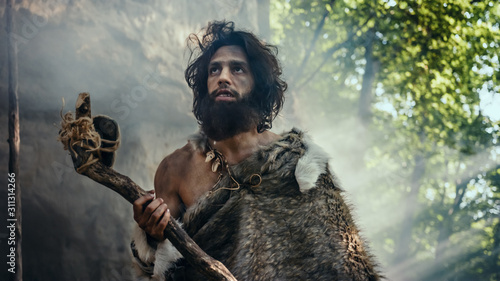 Primeval Caveman Wearing Animal Skin Holds Stone Tipped Hammer Comes out of the Cave and Looks Around Prehistoric Landscape, Ready to Hunt Animal Prey. Neanderthal Going to Hunt in the Jungle