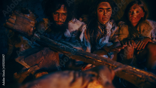 Neanderthal or Homo Sapiens Family Cooking Animal Meat over Bonfire and then Eating it. Tribe of Prehistoric Hunter-Gatherers Wearing Animal Skins Eating in a Dark Scary Cave at Night.