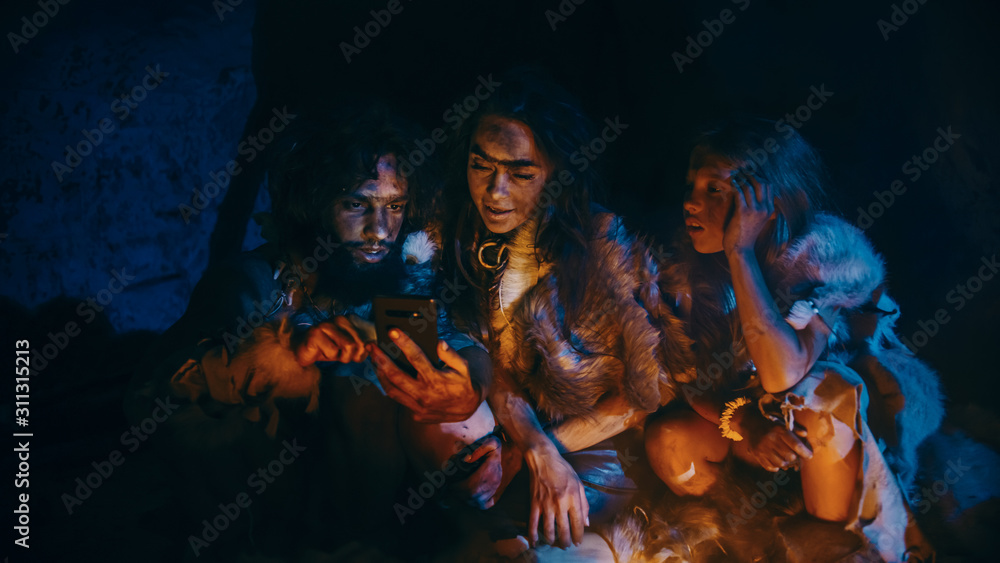 Tribe of Prehistoric, Primitive Hunter-Gatherers Wearing Animal Skins Use Smartphone in a Cave at Night. Neanderthal / Homo Sapiens Family Browsing Internet on Mobile Phone, Cooking Food over Fire