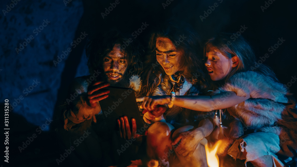 Tribe of Prehistoric, Primitive Hunter-Gatherers Wearing Animal Skins Use Digital Tablet Computer in a Cave at Night. Neanderthal or Homo Sapiens Family Browsing Internet, Watching Videos, TV Shows 