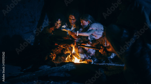 Tribe of Prehistoric, Primitive Hunter-Gatherers Wearing Animal Skins Use Digital Tablet Computer in a Cave at Night. Neanderthal or Homo Sapiens Family Browsing Internet, Watching Videos, TV Shows 