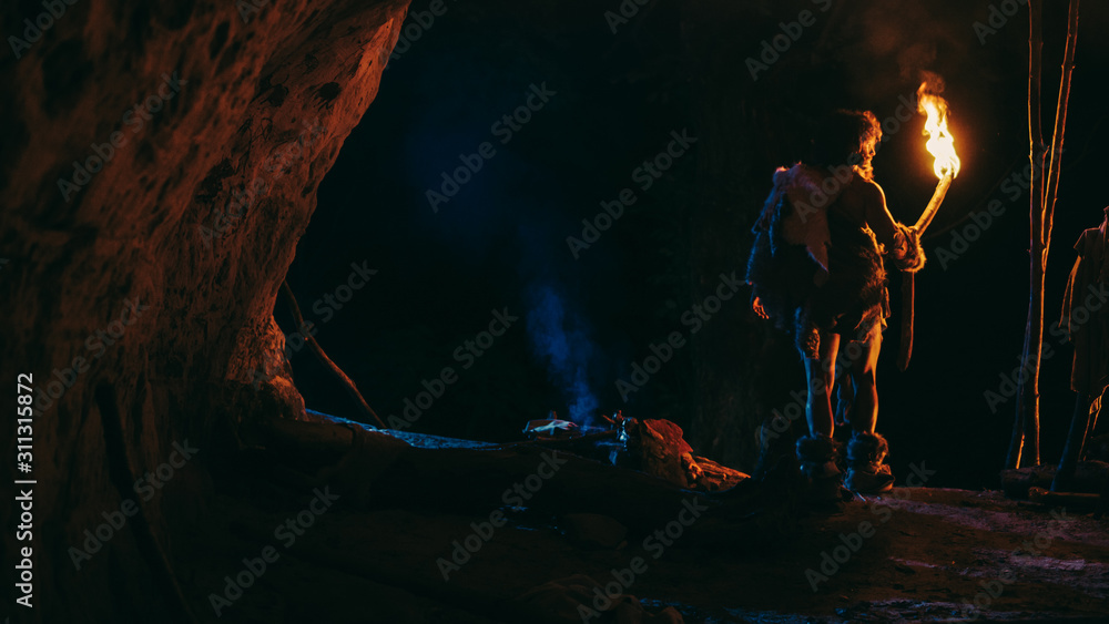 Primeval Caveman Wearing Animal Skin Stands in a Cave At Night, Holding