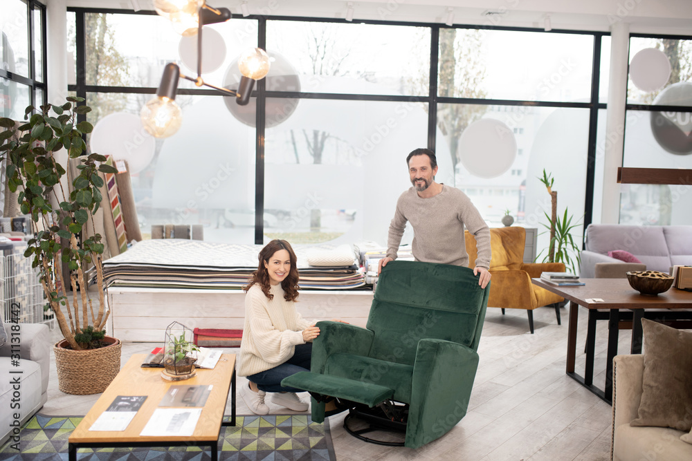 Man and woman in light sweaters looking at big green armchair Man and woman in light sweaters looking at big green armchair