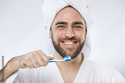 Horizontal studio portrait of happy young man wearing white towel headwrap and bathrobe holding toothbrush looking at camera