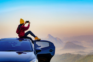young woman sitting on the car roof with enjoy taking photo with mobile phone to the nature of mist in the mountain at sunrise morning, cheerfully life travel