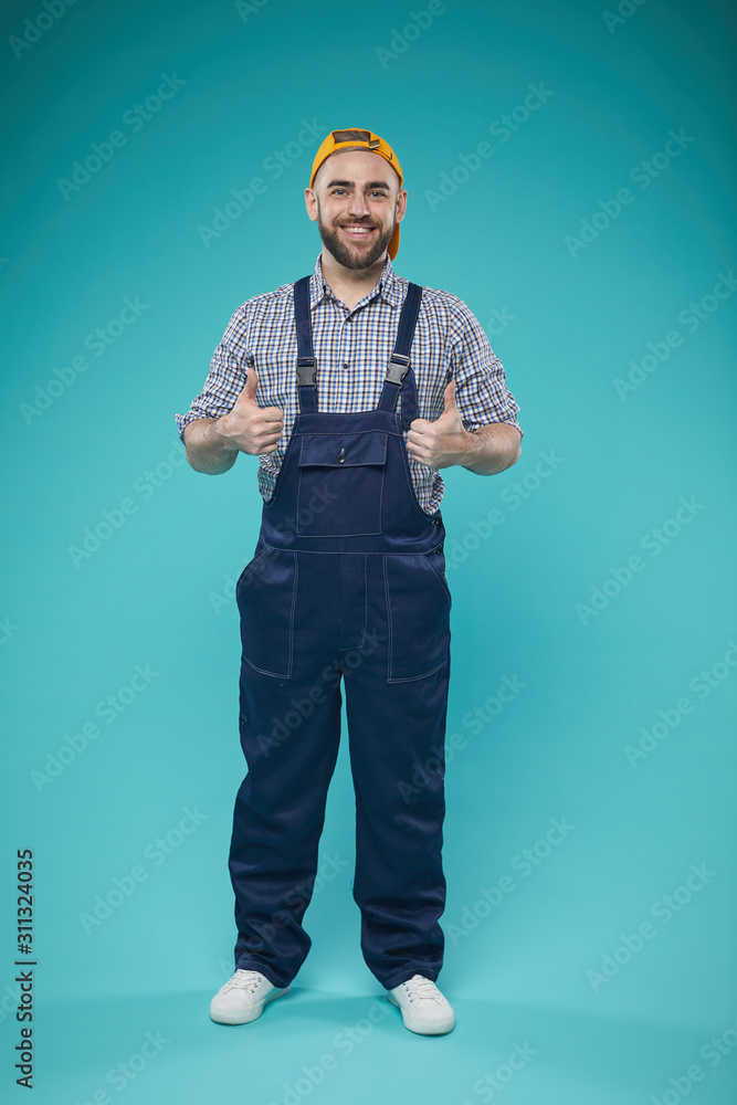 Studio full shot of young adult man standing against turquoise background showing thumbs up looking at camera smiling Studio full shot of young adult man standing against turquoise background showing thumbs up looking at camera smiling