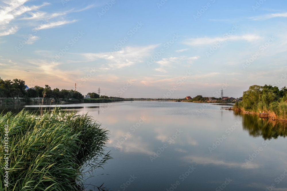 A large river with green reeds on the shore. A blue sky with light clouds reflected in the water.