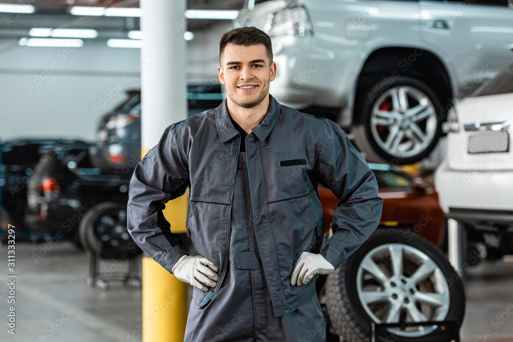 smiling mechanic looking at camera while standing in workshop with hands on hips
