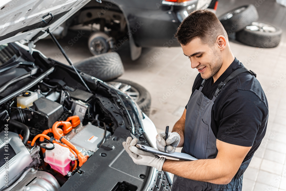 smiling mechanic writing on clipboard while inspecting car engine compartment smiling mechanic writing on clipboard while inspecting car engine compartment