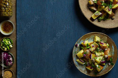 Potato salad with Dijon dressing with ingredients: capers, mustard seeds, red and green onions. Textured blue napkin. Top view, horizontal layout, copy space.