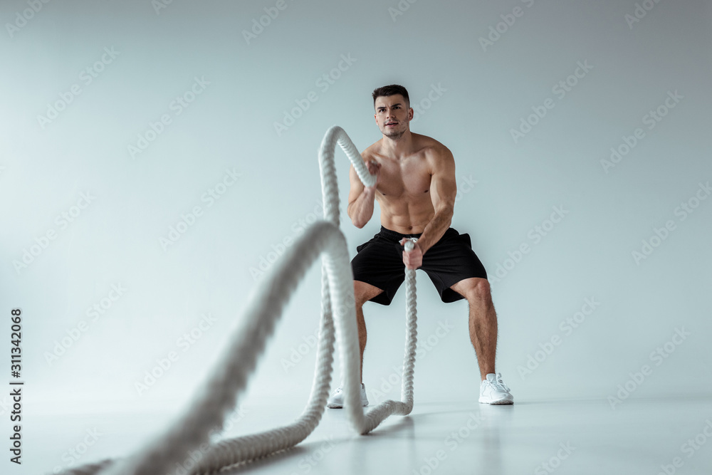 selective focus of sexy muscular bodybuilder with bare torso exercising with battle rope on grey background