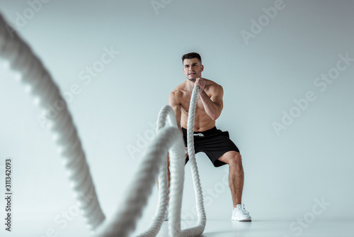 selective focus of sexy muscular bodybuilder with bare torso exercising with battle rope on grey background © LIGHTFIELD STUDIOS