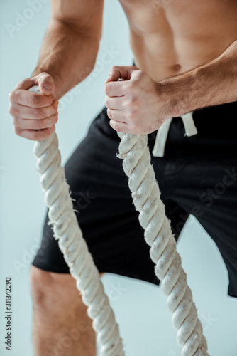 cropped view of sexy muscular bodybuilder with bare torso exercising with battle rope isolated on grey