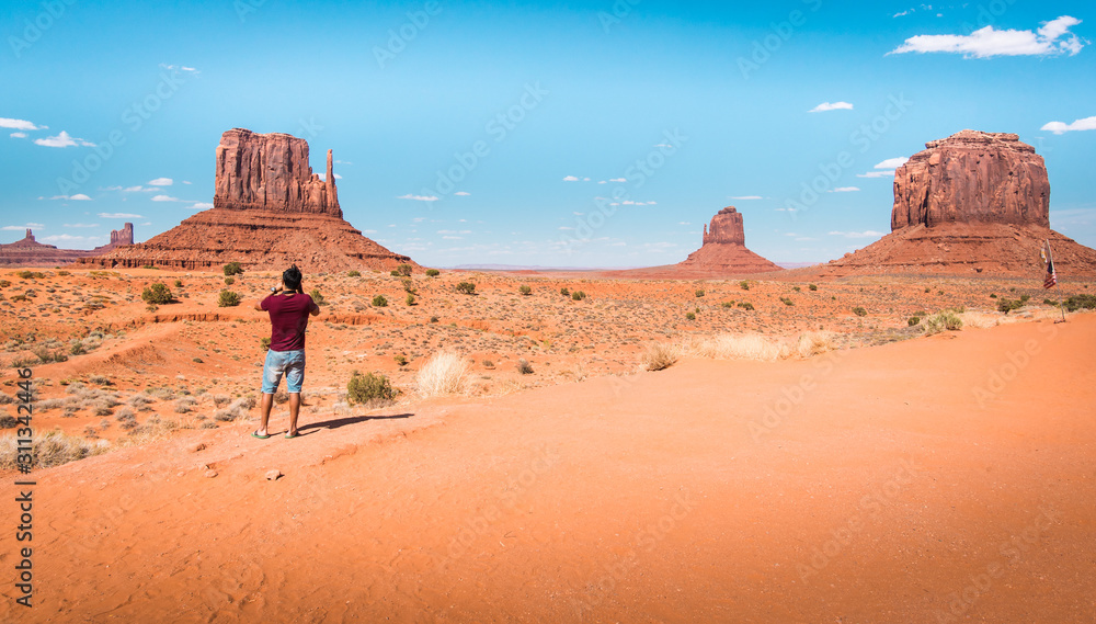 Fototapeta premium Amazed young Latino American traveler in flip flops takes photo of famous Monument Valley desert landscape of large red rock domes on Utah-Arizona border, United States of America