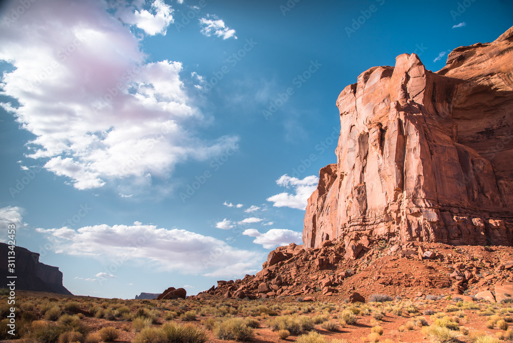 Desert landscape of large sandstone butte, red sand, dry  grass, blue sky and white clouds background. Utah-Arizona border, U.S. of America. Closeup of Elephant Rock taken from Monument Valley floor.