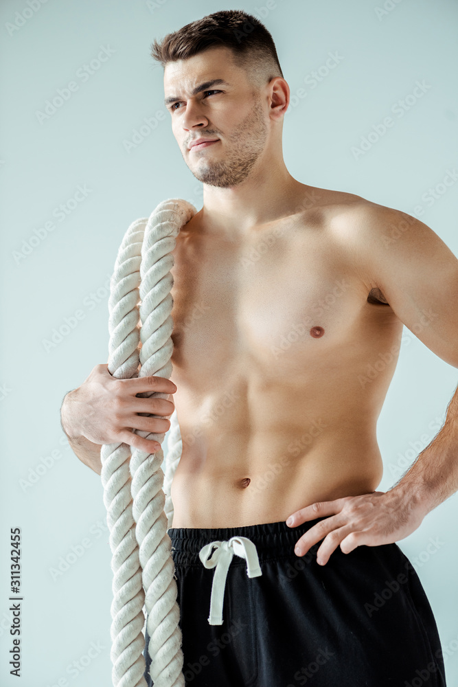 Fototapeta premium niski kąt widzenia sexy muskularny kulturysta z nagim torsem, pozowanie z liny bojowej na szarym tle