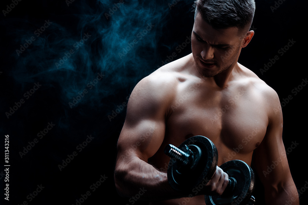 sexy muscular bodybuilder with bare torso excising with dumbbell on black with smoke