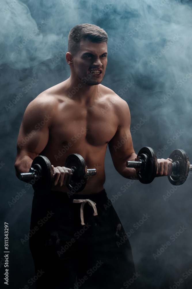 sexy muscular bodybuilder with bare torso excising with dumbbells on black background with smoke