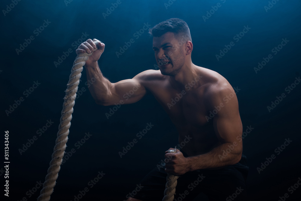 tense muscular bodybuilder with bare torso excising with battle rope on black background with smoke