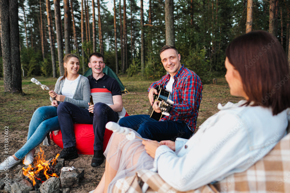 A group of friends relax in a forest camp. Men and women prepare a marshmallow on a bonfire. A party in nature.