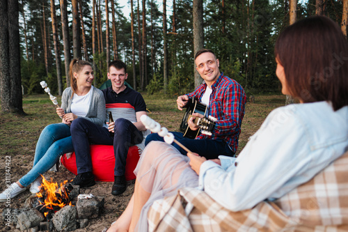 A group of friends relax in a forest camp. Men and women prepare a marshmallow on a bonfire. A party in nature. © spaskov