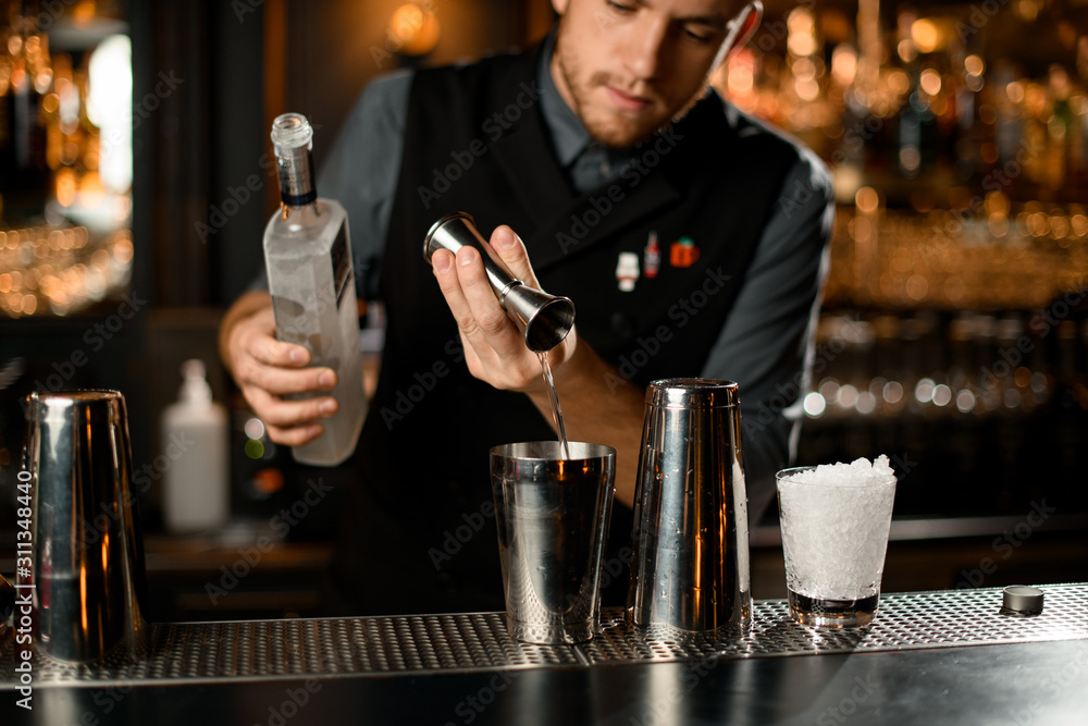 Male bartender pouring a vodka from the jigger to a steel shaker holding a bottle