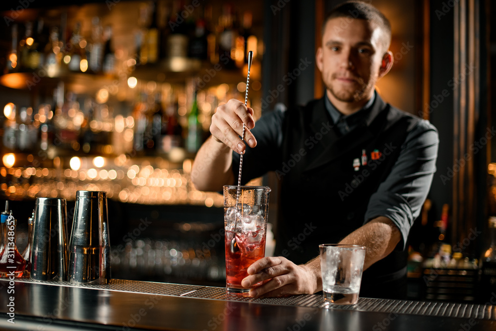 Professional male bartender stirring a red alcoholic drink in the glass with the spoon