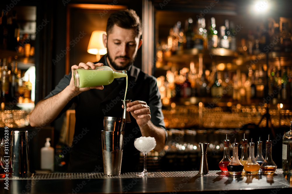 Male bartender pouring a pistachio color liqour from the glass bottle to the steel jigger