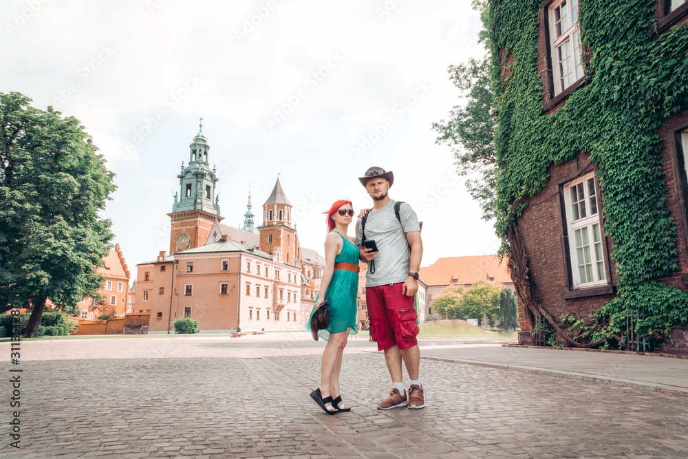 Young couple in love is standing on the street near the Wavel castle in Krakow, Poland. Tourism travel concept, copy space. Man and woman, summer day. leather hat and sunglasses, smartphone in hand.