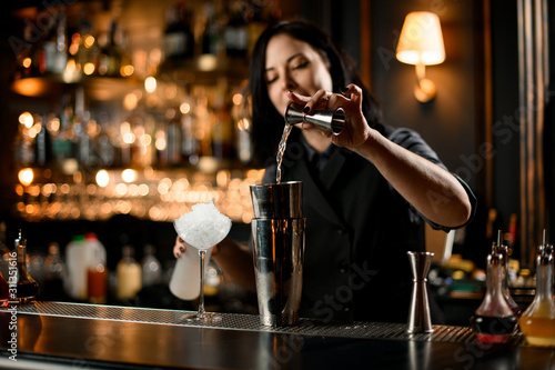 Bartender girl pouring an alcoholic drink from the jigger to a professional steel shaker photo