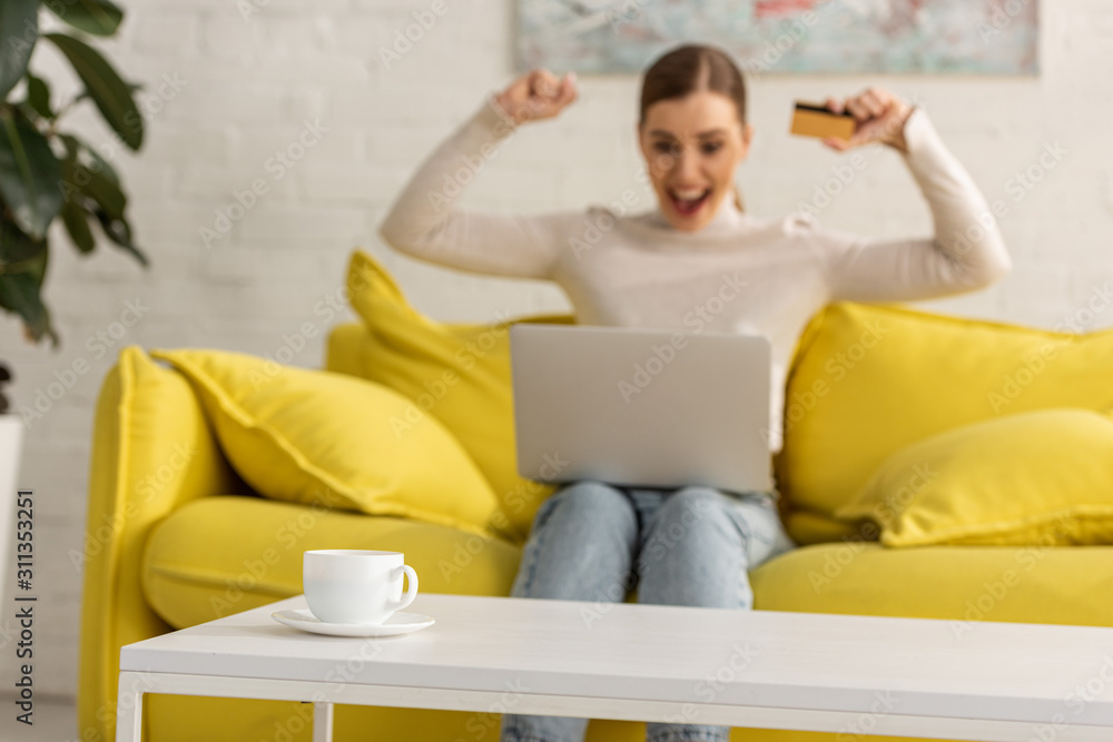 Selective focus of coffee on table and excited woman with credit card and laptop on sofa at background Selective focus of coffee on table and excited woman with credit card and laptop on sofa at background