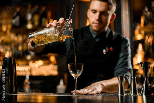 Professional bartender pouring yellow transparent alcoholic drink from the measuring cup to the glass through the strainer filter