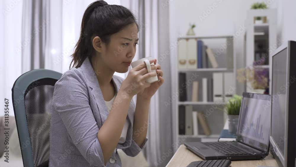 Focused side view young woman frowning and typing on desktop computer keyboard while working in modern home office. young asian japanese lady in suit thinking solution and drinking cup of coffee. Focused side view young woman frowning and typing on desktop computer keyboard while working in modern home office. young asian japanese lady in suit thinking solution and drinking cup of coffee.