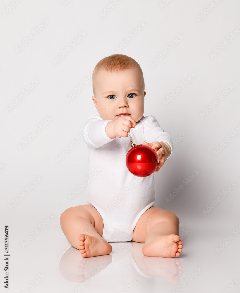 Infant baby blond boy toddler in white bodysuit sitting holding handed playing with red Christmas tree red ball on white Infant baby blond boy toddler in white bodysuit sitting holding handed playing with red Christmas tree red ball on white