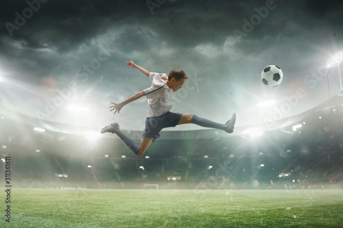 Faster than wind. Young male football or soccer player in sportwear and boots kicking ball for the goal on stadium with flashlights on background. Concept of healthy lifestyle, professional sport.