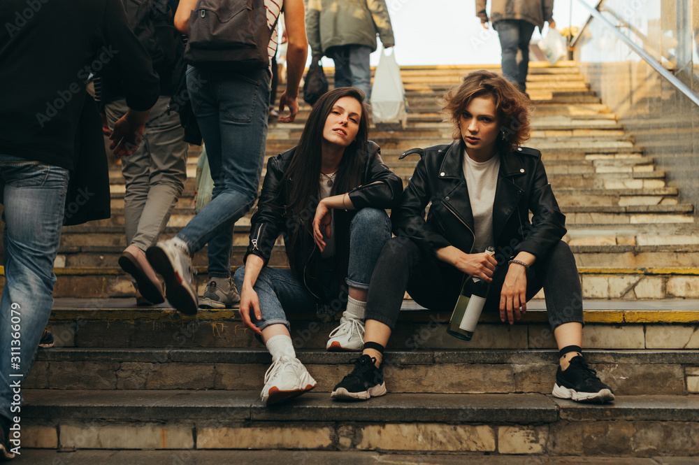 Portrait of two free girls in stylish clothes sitting on the stairs with a bottle of wine in hand in the middle of a crowd of people. Stylish models posing at the exit of subway,sitting on the stairs
