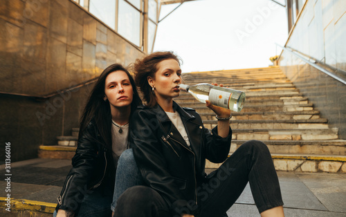 Two girlfriends sitting on the stairs in underground drinking wine from a bottle and posing on camera wearing stylish casual clothes.Girls in leather jackets sitting in the underground on the stairs