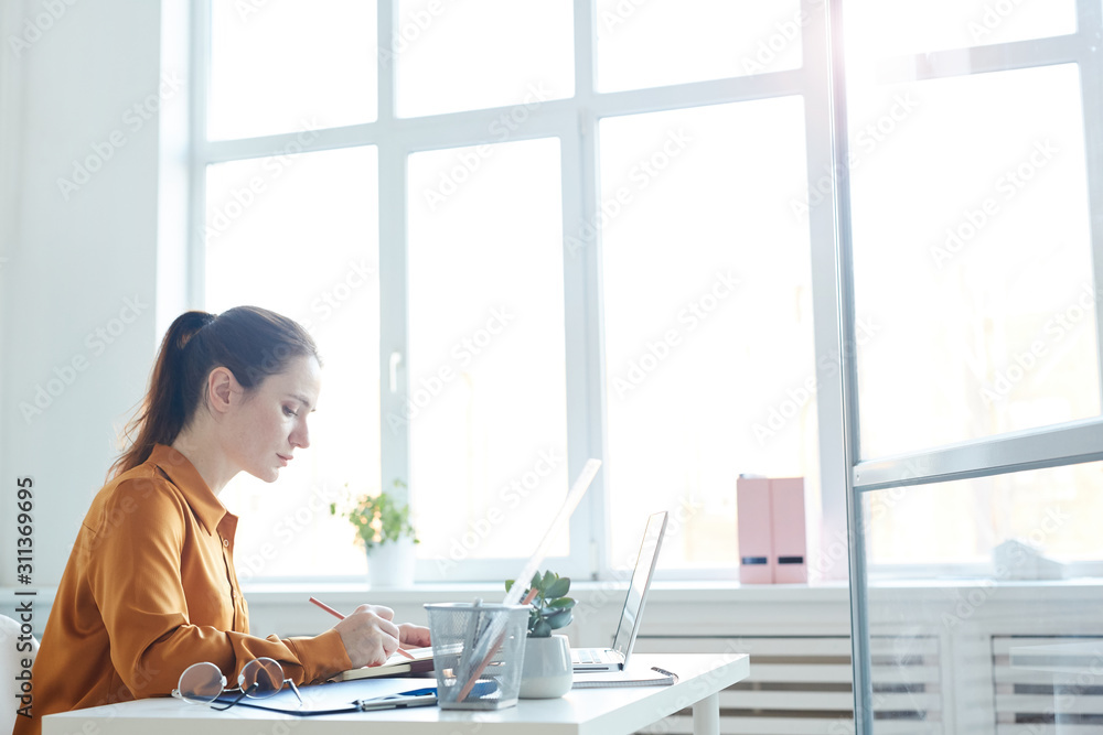 Young serious manager sitting at her workplace in front of laptop and writing in documents she working with report at office Young serious manager sitting at her workplace in front of laptop and writing in documents she working with report at office