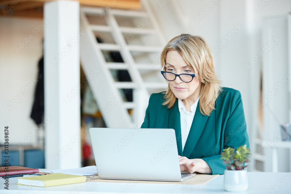 Serious mature manager in eyeglasses sitting at the table and typing on laptop computer she working online