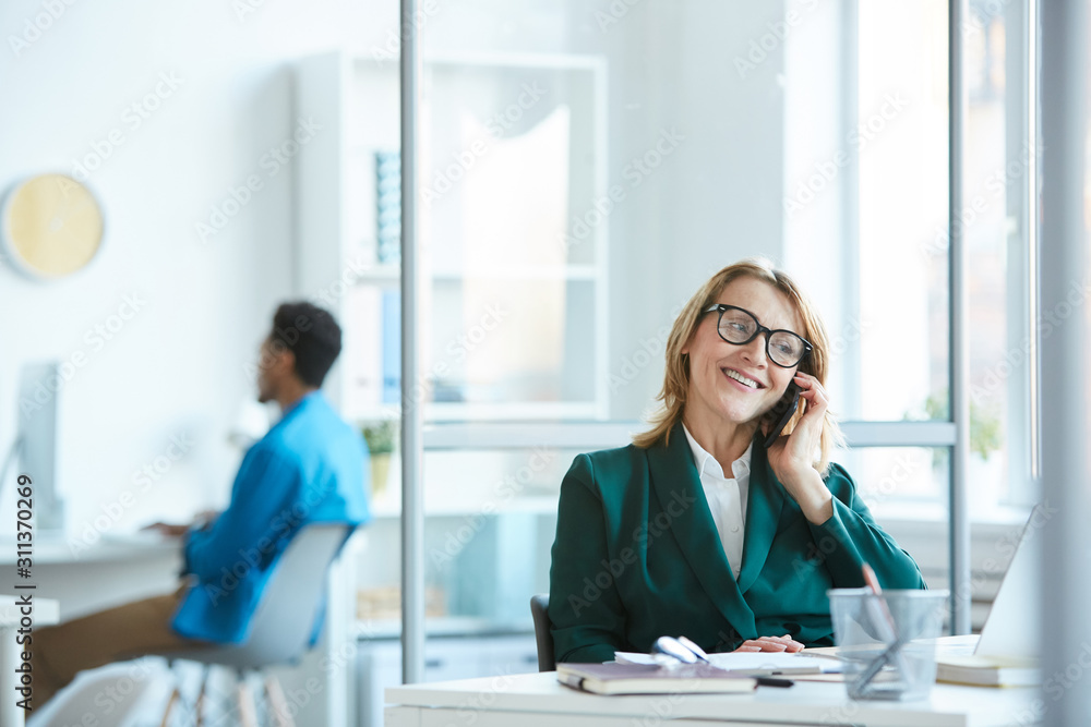Mature manager in eyeglasses sitting at the table talking on mobile phone and smiling at office Mature manager in eyeglasses sitting at the table talking on mobile phone and smiling at office