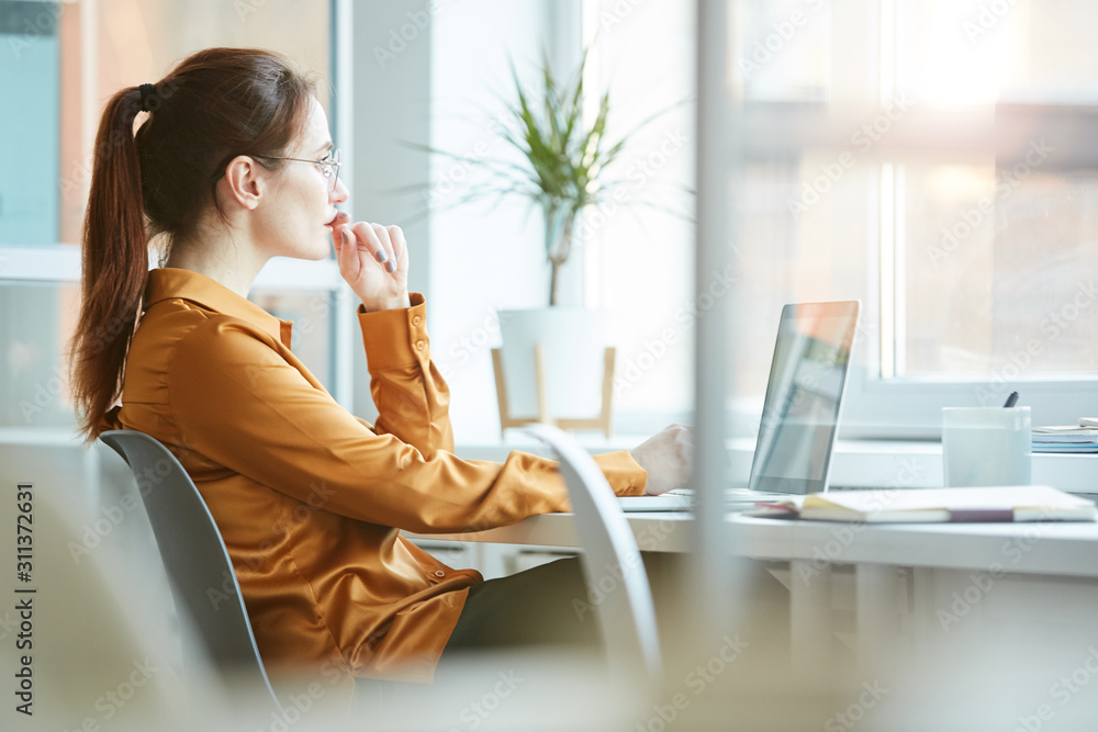 Young woman sitting at her workplace in front of the window with pensive sight and thinking about something while working on laptop