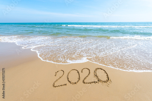Text 2020 Happy New Year on the beach with waves and clear blue sea. 2020 numbers on the coast. Hand written messages in sand on a beautiful beach background