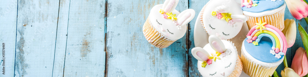 Easter holiday greeting card background. Cute homemade cupcakes with traditional Easter bunny, egg and springtime flowers decor. Happy easter concept. Copy space for your text - obrazy, fototapety, plakaty 