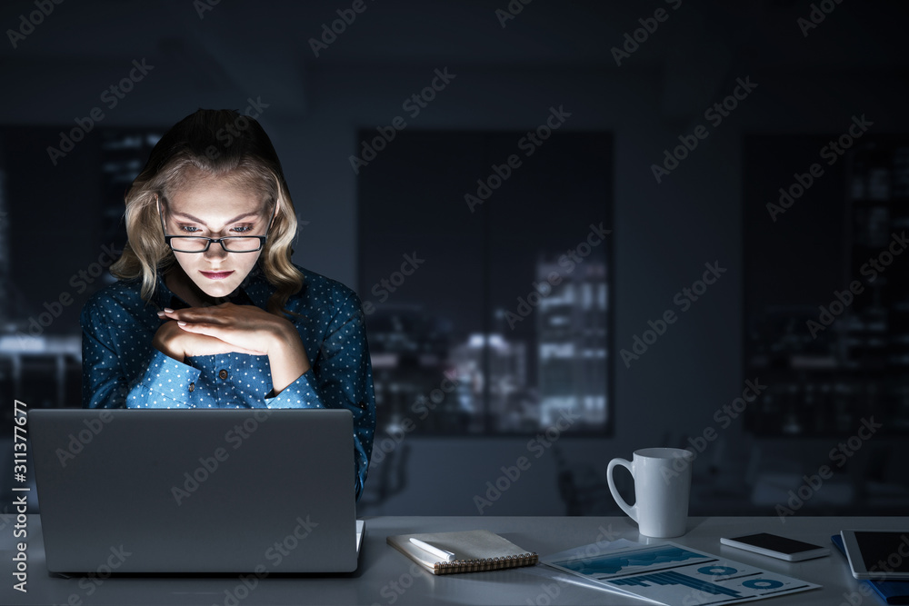 Attractive blonde working on laptop in dark office. Mixed media