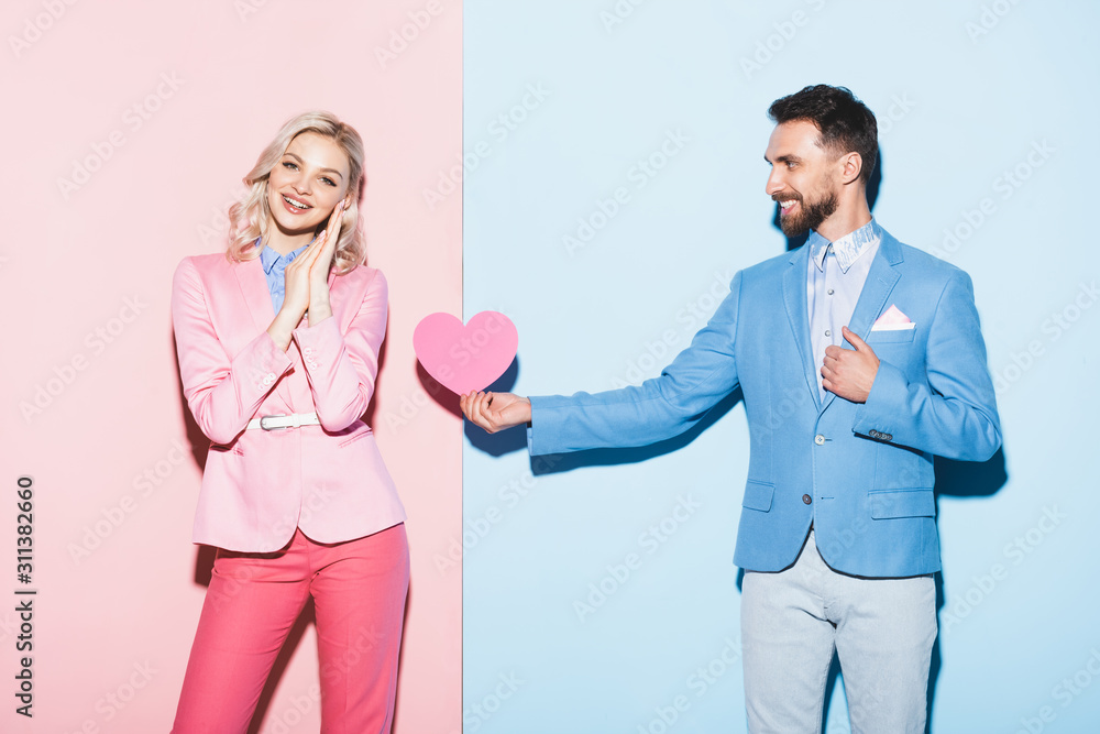 handsome man giving heart-shaped card to smiling woman on pink and blue background