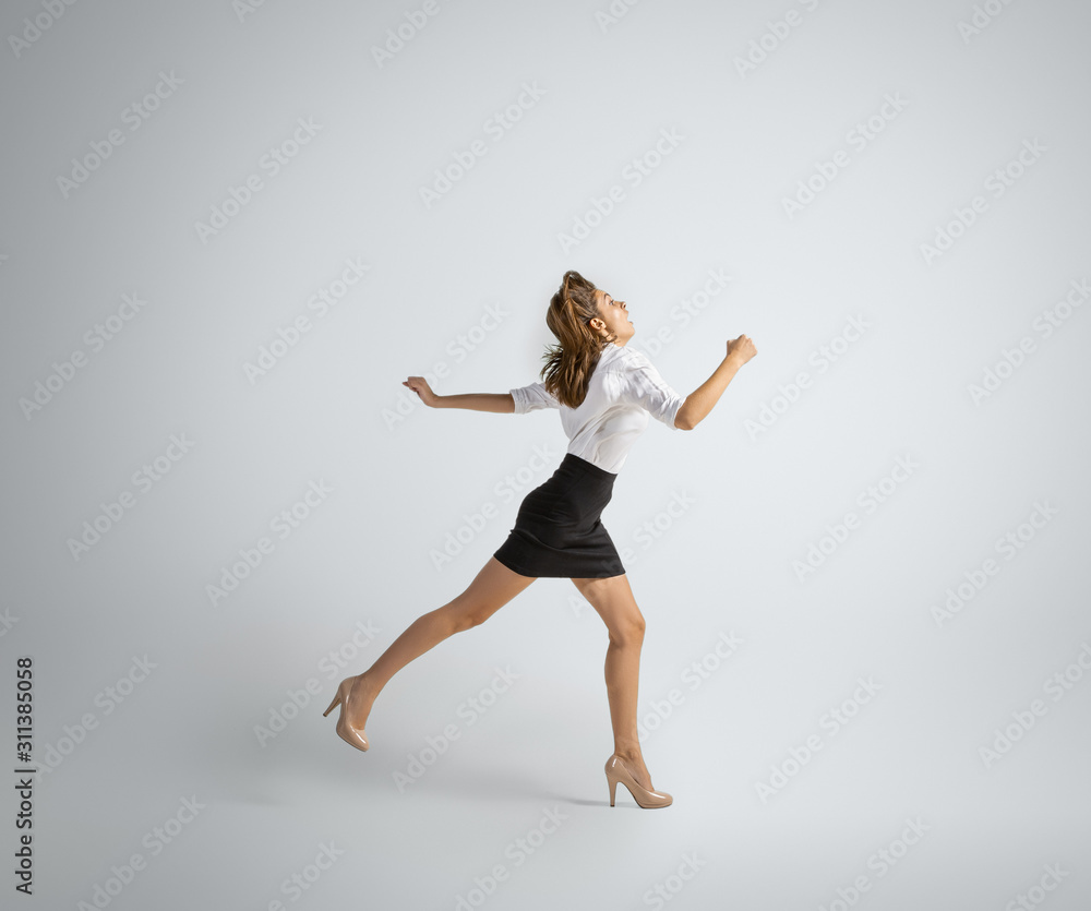 Obraz premium Hurrying up to new goals. Woman in office clothes running on grey studio background. Businesswoman training in motion, action. Unusual look for sport, new activity. Sport, healthy lifestyle.