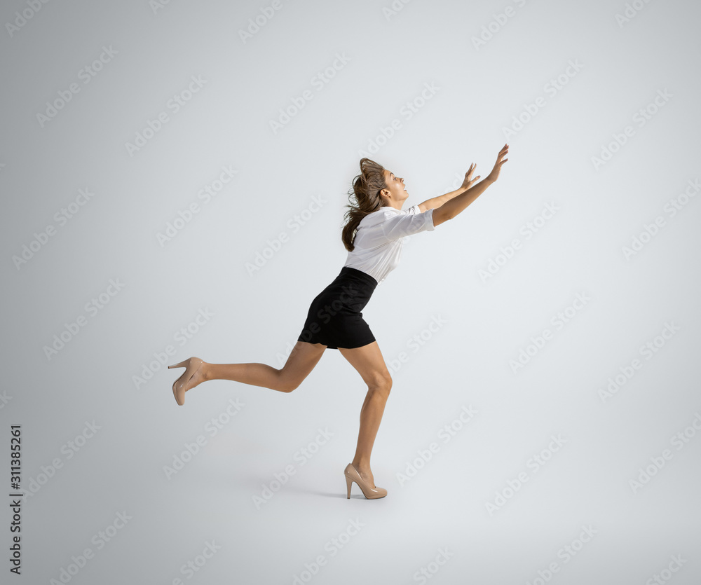 Obraz premium Hurrying up to new goals. Woman in office clothes running on grey studio background. Businesswoman training in motion, action. Unusual look for sport, new activity. Sport, healthy lifestyle.