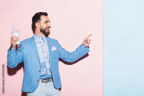  smiling man holding cocktail and pointing with finger on pink and blue background